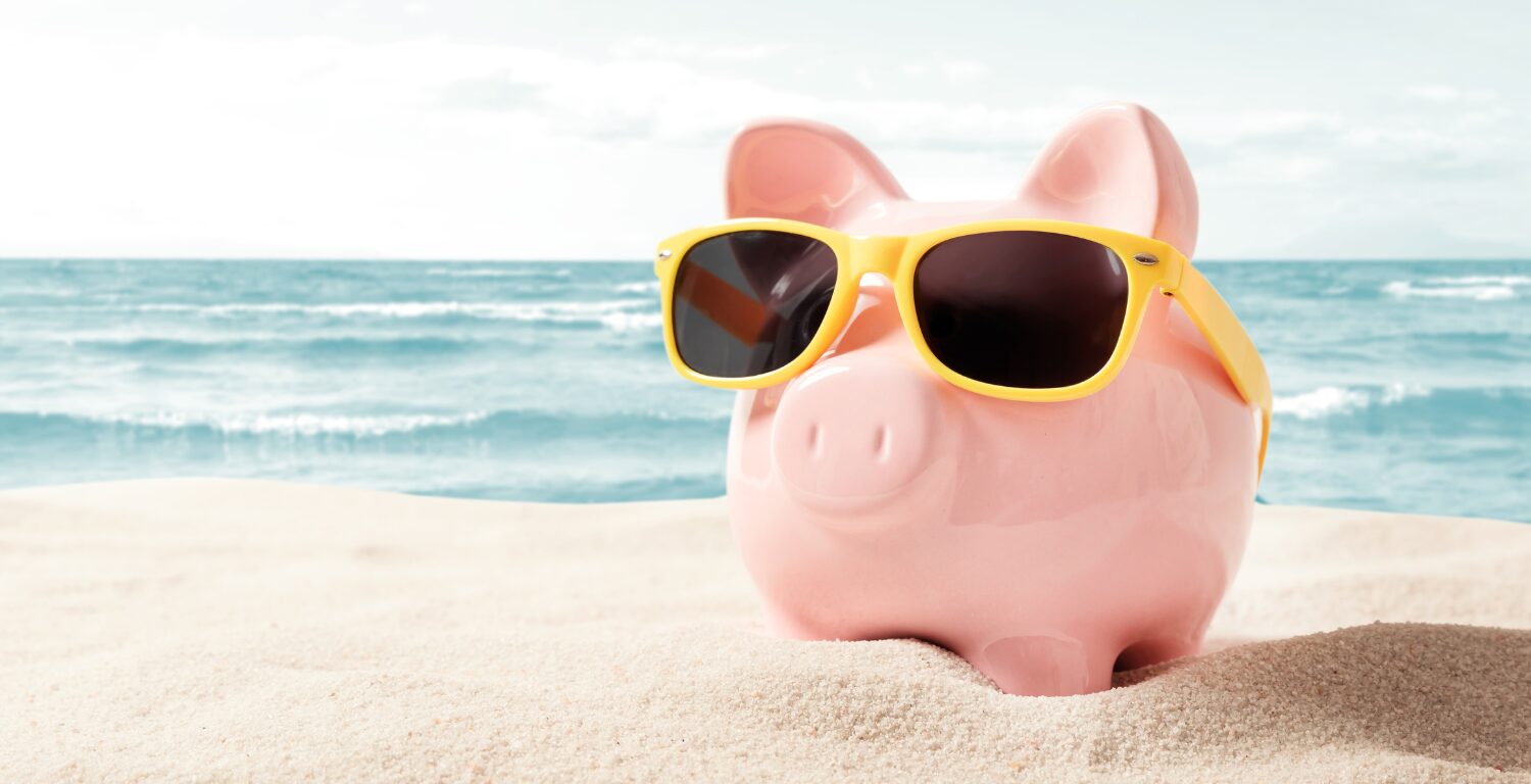 piggy bank in yellow sunglasses relaxing on a white sand beach with the ocean in the background