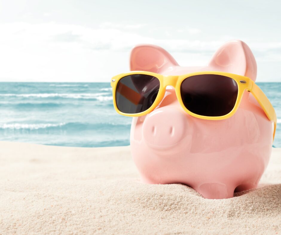 piggy bank in yellow sunglasses relaxing on a white sand beach with the ocean in the background